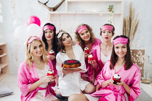 Group of pretty and lovely bridesmaids in pink robes with beautiful bride holding cupcakes and cupcakes in light room. Air balloons in background.