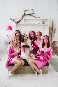 Group of pretty and lovely bridesmaids in pink robes with beautiful bride holding cupcakes and cupcakes in light room. Air balloons in background.