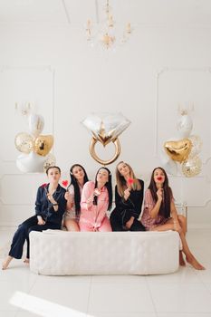 Stock photo of five girls in pajamas eating sweet lollipops sitting close on big white bed in beautiful white room with inflated air balloons in shape of wedding ring, hearts and stars.