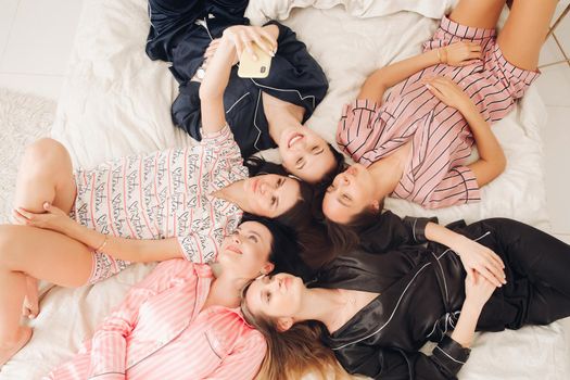 Top view stock photo of five beautiful girlfriends in lovely home apparel making selfie laying on cozy bed. Bride-to-be taking selfie with her mobile phone with her best friends or bridesmaids while resting on bed. They are making duck faces posing to camera.