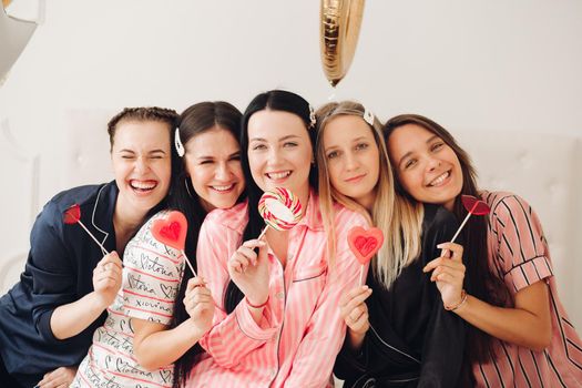 Stock photo portrait of lovely beautiful adult girls smiling at camera holding sweet lollipops in front of their lips. Bachelorette party. Hen party concept. Bridesmaids friends and the bride in the middle with lollipops.