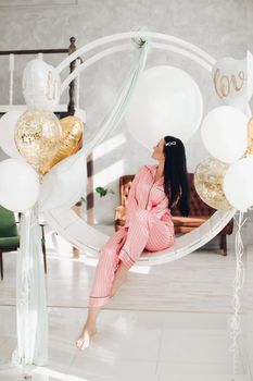 Studio portrait of beautiful young black-haired girl in pink striped pyjamas sitting elegantly on hanging roung chair surrounded by white air balloons.