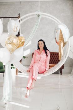 Studio portrait of beautiful young black-haired girl in pink striped pyjamas sitting elegantly on hanging roung chair surrounded by white air balloons.