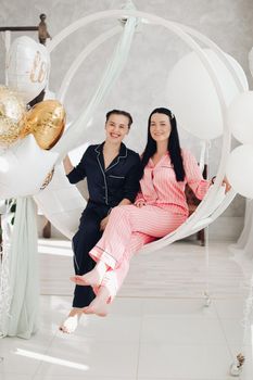 Full length of two beautiful girls in pink striped pyjamas sitting barefoot snuggling on white swings decorated with air balloons. Bride-to-be concept. Hen party with the best friend.