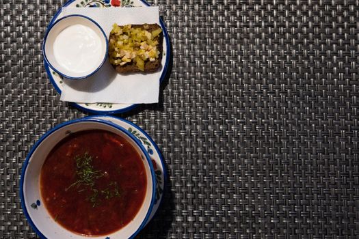 Borscht in a plate is decorated with greens. a cup with sour cream and a sandwich with pickles and bacon for borscht. Plates with napkins decorated with ornaments in a Russian cuisine restaurant.....