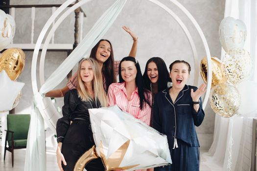 Five friends celebrating the party dedicated to a bachelorette in the middle. Hen party celebration. Girls laughing and smiling with black-haired girl in the middle holding a big balloon ring.