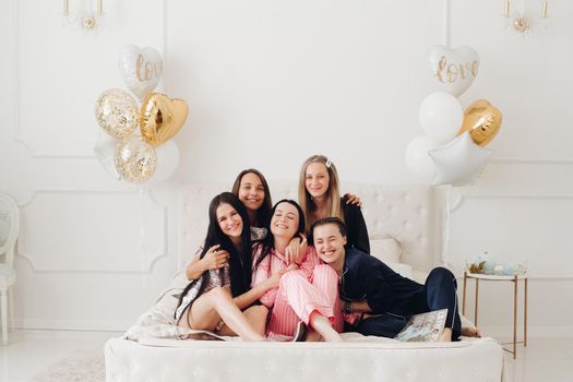 Group of happy girl friends laughing posing on bed enjoying pajamas party full shot. Smiling female in domestic clothing enjoy friendship hugging together having fun celebrating holiday