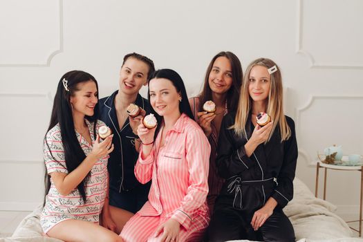 Happy pretty women in sleepwear sitting on the edge of the bed and holding cupcakes. Friendship and happiness concept