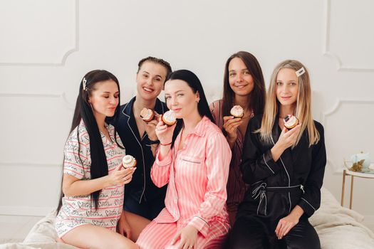 Stock photo of pretty bridesmaids in pyjamas holding delicious handmade cupcakes in front of their lips looking at camera. Celebrating hen party together. Best friends.