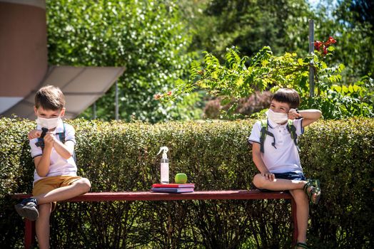 Coronavirus school reopening concept: a school boys sit on bench in front of a school wearing a face mask. Communicating trough smart watch. Covid-19, health, education, safety, and back to school.