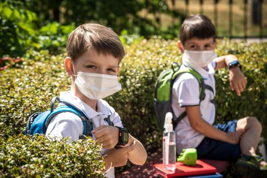 Coronavirus school reopening concept: a school boys sit on bench in front of a school wearing a face mask. Communicating trough smart watch. Covid-19, health, education, safety, and back to school.