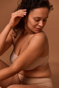 Portrait of a dark-haired curly woman with skin flaws, cellulite and stretch marks posing in beige underwear, looking down against colored background with copy space. Body positive concept