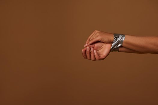 Cropped view of female hands tied with duct tape against a dark beige background, Copy space. Social concept for International Day to End Aggression, Inequality and eliminating violence against Women