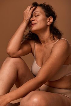 Serene middle aged pretty woman with imperfect body, cellulite and stretch marks wearing beige underwear hiding half of her face posing on colored background with copy space. Body positivity concept