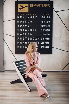 Full length photo of gorgeous woman with long blonde hair in pink suit sitting on modern chair against flights timetable. Waiting at the airport.