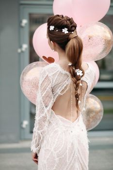 Back view of unrecognizable gorgeous brunette with beautiful hairdo and flowers in it posing in luxurious white dress with bunch on pink air balloons.
