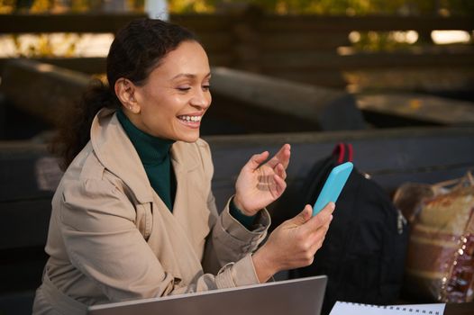 Cheerful attractive woman smiles while making a video call via a mobile app on her smartphone. Young confident business woman working outdoors away from urban civilization on the oak grove autumn cafe