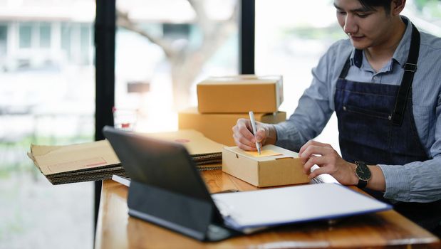 Young entrepreneur SME freelance man using smartphone receive order client and take note working with packaging sort box delivery online market.