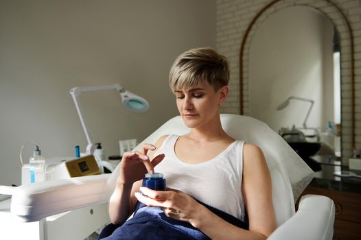 Beautiful young woman sitting on a chair at cosmetology room, holding a bottle with beauty product and applying moisturizer for skin caring