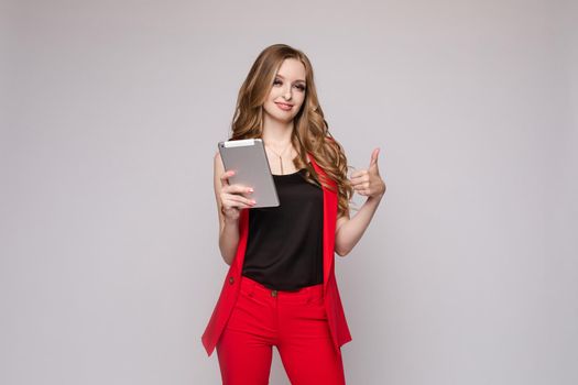 Young beautiful girl holding silver tablet and reading book on it. Brunette lady in red jacket and pants posing with electronic device. Elegant business woman in costume seriously checking email.