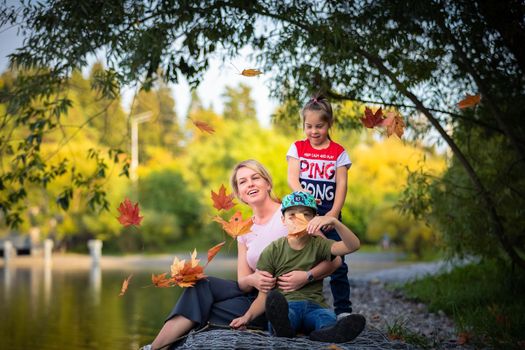 Mom with children is sitting by the pond in the park, autumn leaves are falling from above. Russia Moscow September 24, 2020.