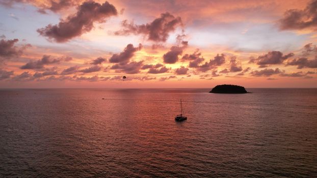 Parasailing at an orange sunset on the beach. Parachute flight in the midst of an epic sunset. Small waves, people relax on the beach, palm trees grow. There is a small island in the distance. Tropics