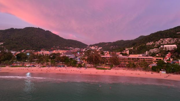 Orange sunset with a view of the beach. People are resting, seeing off the sun over the horizon. Small waves. Clean sand. Hills, clouds and the city are visible in the distance. Dogs are running. Kata