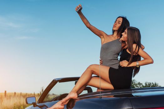 Beautiful two girls are photographed on the road against a background of blue sky and field on a black convertible.