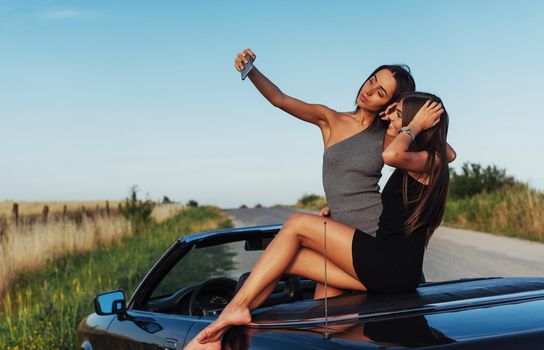 Beautiful two girls are photographed on the road against a background of blue sky and field on a black convertible.