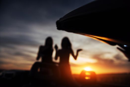 Two girls happy to pose next to a black car against the sky on a fantastic sunset. Natural blurred background. Soft light effect.