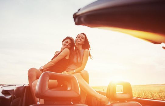 Young two women at a photo shoot. Girls gladly posing next to a black car against the sky on a fantastic sunset.