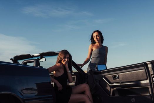 Beautiful two girls are photographed on the road against a background of blue sky and field on a black convertible.