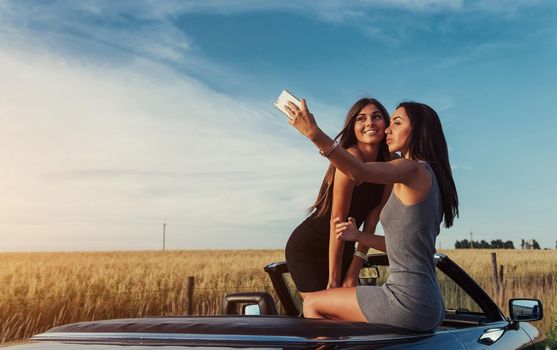 Beautiful two girls are photographed on the road against a background of blue sky and field on a black convertible.