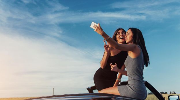 Beautiful two girls are photographed on the road against a background of blue sky and field on a black convertible.