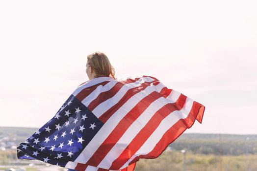 Young beautiful woman holding USA flag. The back of a young woman with the US national flag on her shoulders against the backdrop of the mountains celebrates the Independence Day of the United States.