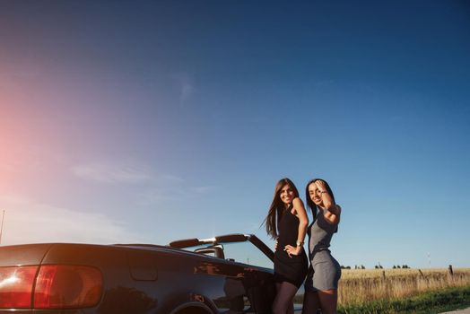 Young women at a photo shoot. Girls gladly posing next to a black convertible against the sky. Summer walk.