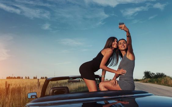 Beautiful two girls are photographed on the road against a background of blue sky and field on a black convertible.