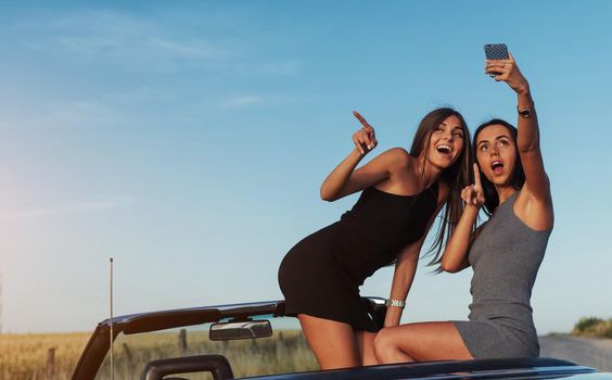 Beautiful two girls are photographed on the road against a background of blue sky and field on a black convertible.