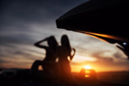 Two girls happy to pose next to a black car against the sky on a fantastic sunset. Natural blurred background. Soft light effect.