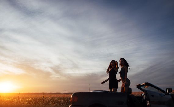 Young two women at a photo shoot. Girls gladly posing next to a black car against the sky on a fantastic sunset.