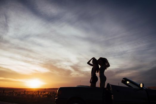 Young two women at a photo shoot. Girls gladly posing next to a black car against the sky on a fantastic sunset.