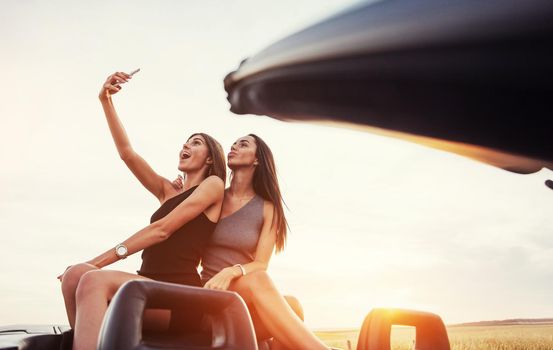 Young two women at a photo shoot. Girls gladly posing next to a black car against the sky on a fantastic sunset.