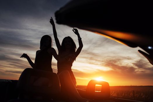 Young two women at a photo shoot. Girls gladly posing next to a black car against the sky on a fantastic sunset.