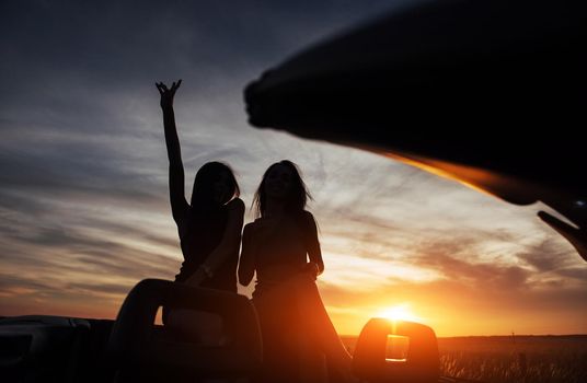 Young two women at a photo shoot. Girls gladly posing next to a black car against the sky on a fantastic sunset.