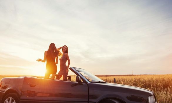 Young two women at a photo shoot. Girls gladly posing next to a black car against the sky on a fantastic sunset.
