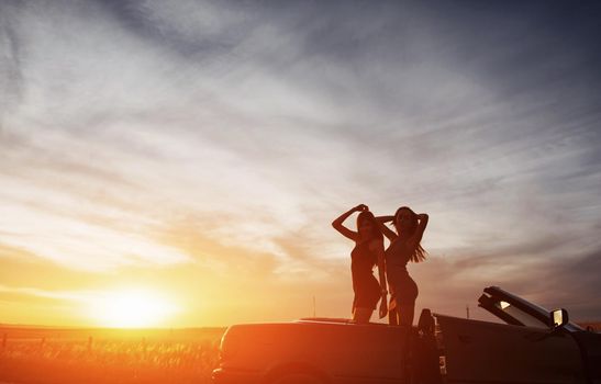 Young two women at a photo shoot. Girls gladly posing next to a black car against the sky on a fantastic sunset.