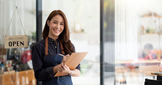 Portrait of a smiling Asian entrepreneur standing behind her cafe counter with open sign board.