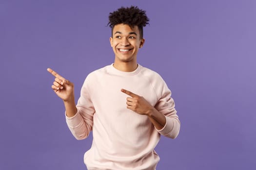 Portrait of enthusiastic, good-looking hispanic man with dreads, looking and pointing upper left corner with satisfied beaming smile, seeing cool thing, made his choice, purple background.