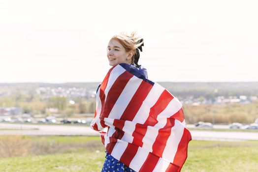 A cute young woman is wrapped in a usa flag and looks out over the city. 4th of July.