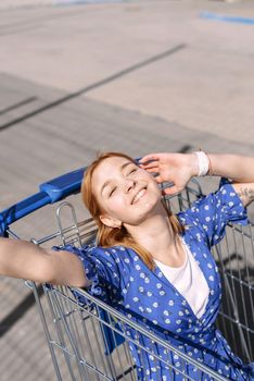 Cheerful stylish young woman in a blue dress on a sunny day sits in a shopping trolley in front of a supermarket. Fashion cool smiling girl.